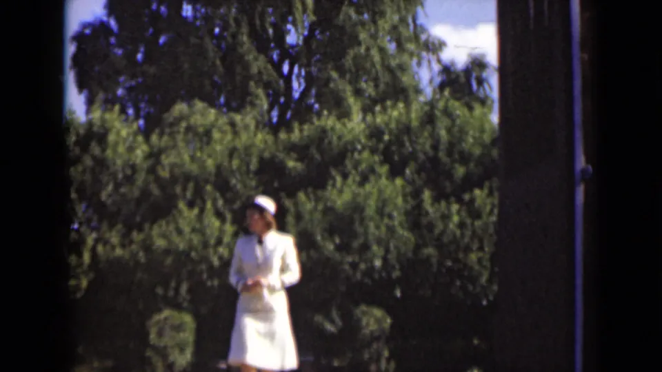 A woman in a white nurse-style uniform stands on a garden path, with hedges, trees, and a vertical post visible in the frame.