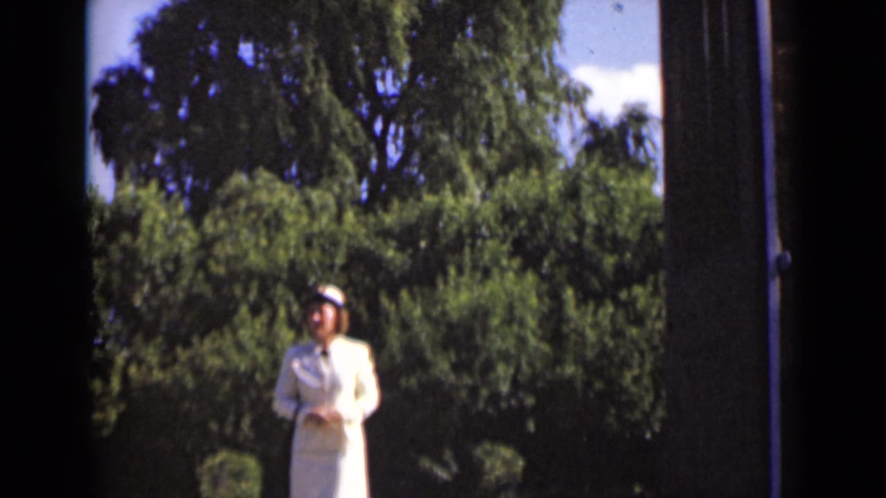 Woman in a white suit and hat stands outside beyond a dark open doorway with trees behind her.