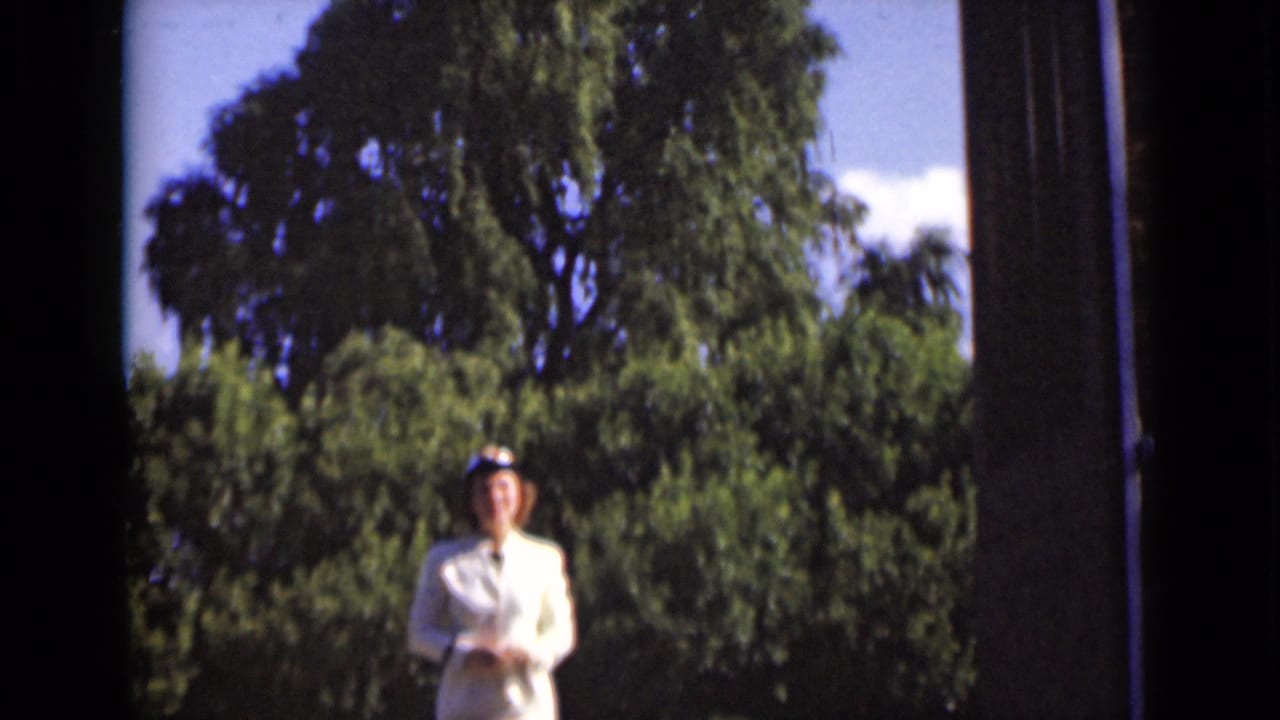 Woman in a white suit faces the camera outside, framed by the doorway and green trees.