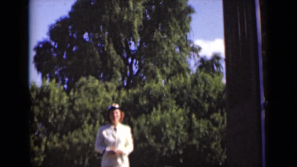 The woman in a white suit and hat faces the camera from the sunny exterior, framed by the open doorway and trees in New York footage.