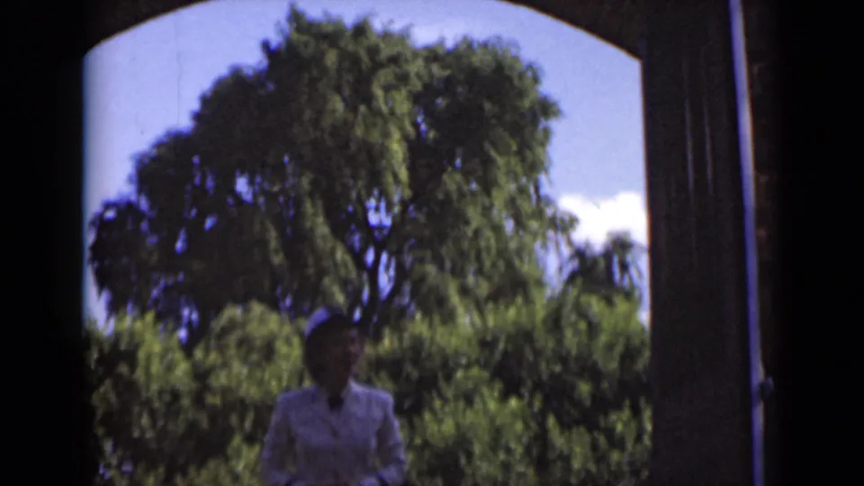 Near the end of the clip, the woman in a white suit is partly shadowed below the doorway opening, with dense green trees and sky behind her.