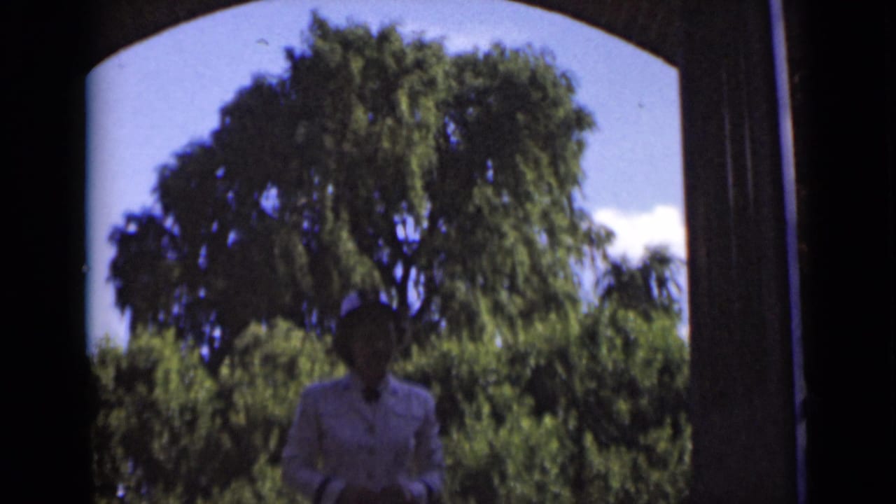 A woman in a white dress and hat appears under a shaded archway in 1947 New York home-movie footage, with leafy trees and blue sky beyond.