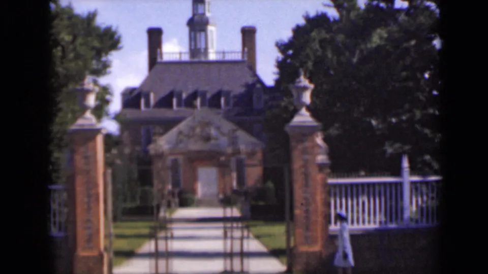 A gated walkway opens toward a stately brick building with a cupola, white fencing, and mature trees in 1947 New York home-movie footage.