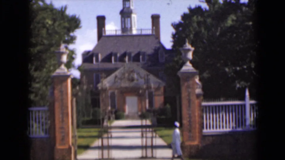 The woman in white moves along the gated approach toward the brick building, framed by trees, white fencing, and bright daylight in New York.