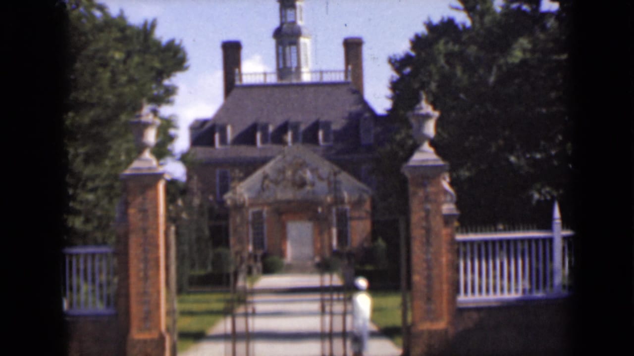 Woman in light clothing walking through a brick gate toward a large house.