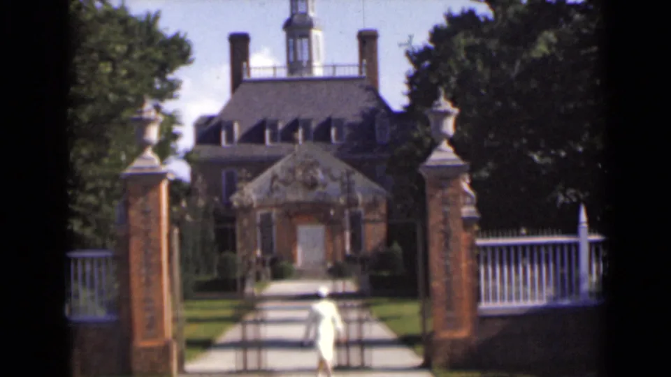 The woman moves farther inside the gated approach, with lawn, trees, white fence, and a cupola-topped house beyond.