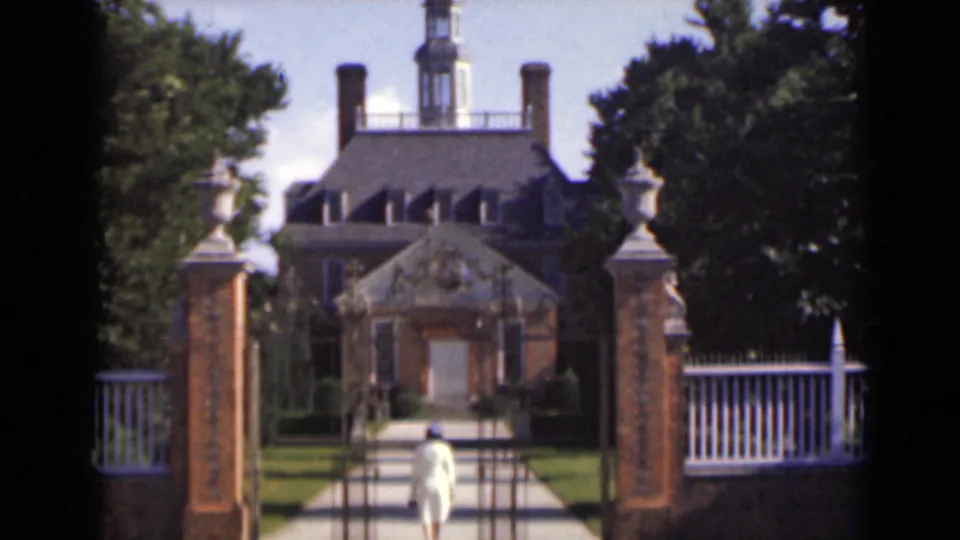 A rear view shows the woman on the central path inside the gate, framed by brick posts, fence rails, lawn, and the house facade.