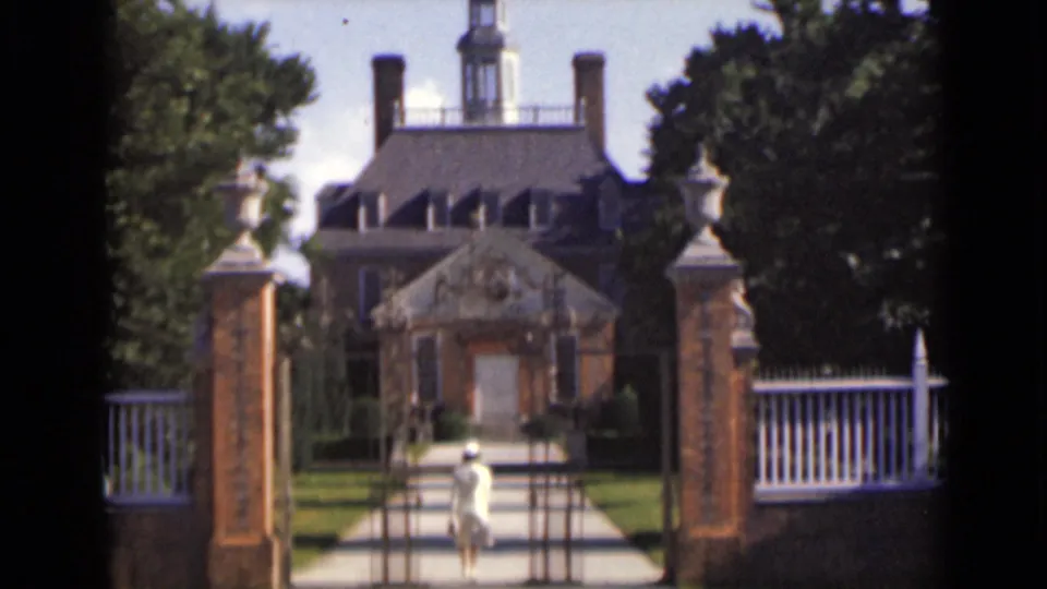 At the end of the clip, the woman remains on the estate walkway as the large house, cupola, chimneys, trees, and white fence stay in view.