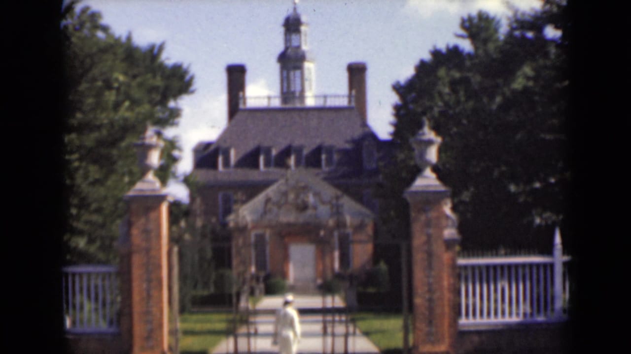 Woman in white walking through a gated path toward a large brick mansion.
