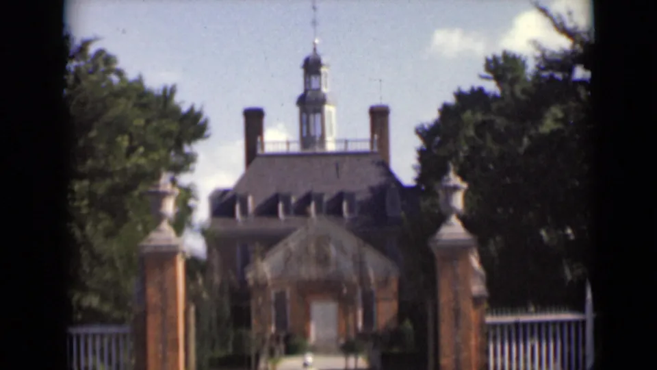 Brick colonial-style mansion in New York with cupola, chimneys, gateposts, white fencing, and trees in soft 8mm color.