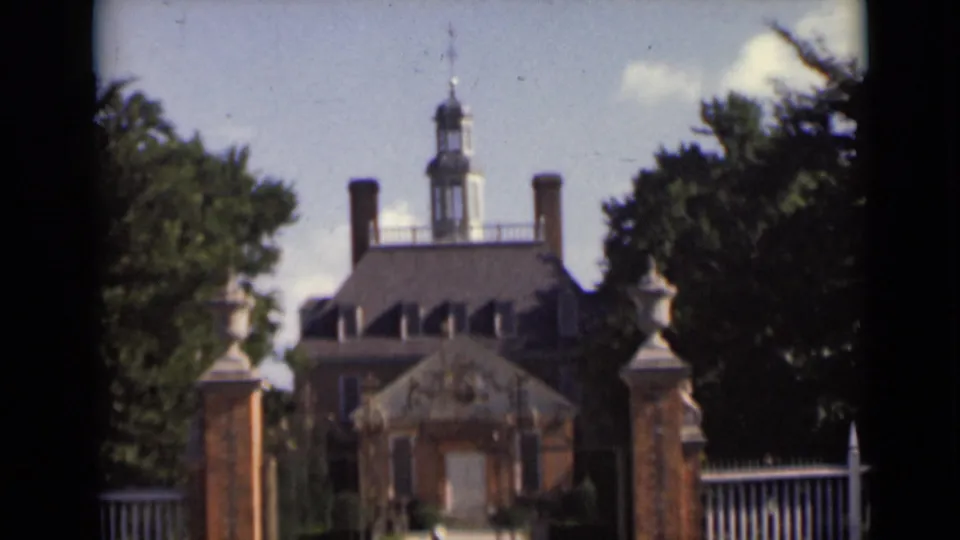 Woman in white walking closer to a brick mansion entrance, with a cupola, chimneys, gateposts, and trees visible.
