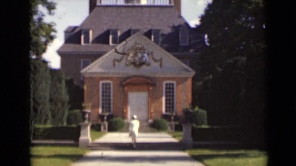 Closer view of a woman in white nearing the front steps of a large brick mansion with decorative pediment and hedges.