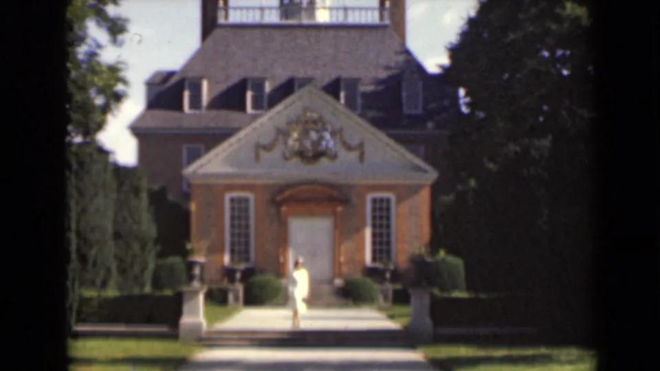 A woman approaches the entrance of a formal brick building in 1947 New York, framed by hedges, trees, and gold-toned ornament.