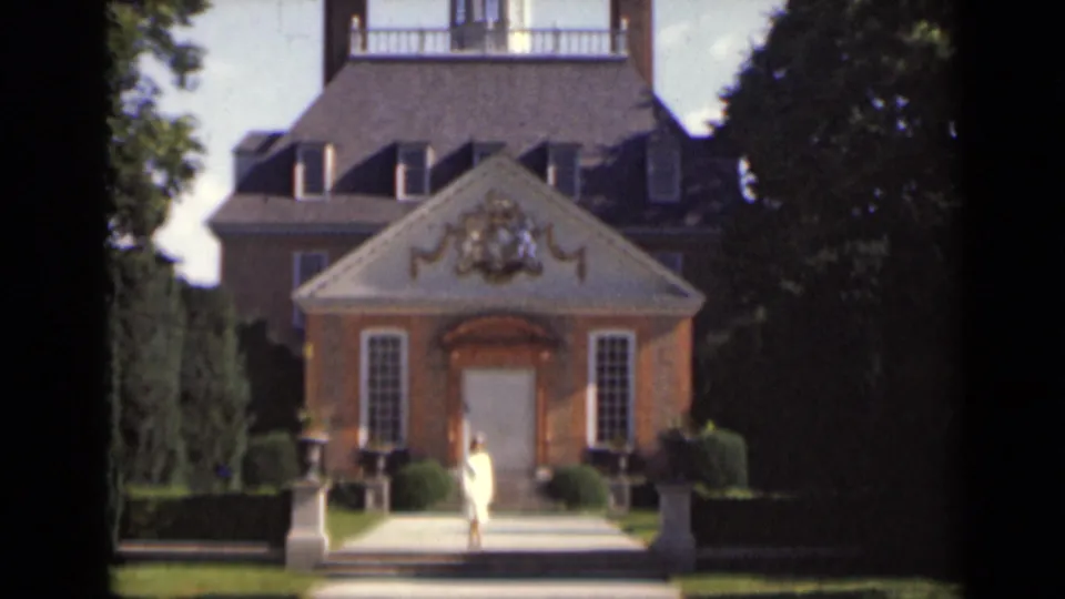 The woman pauses near the steps of the ornate building exterior, with a decorated pediment, doorway, hedges, and trees visible.