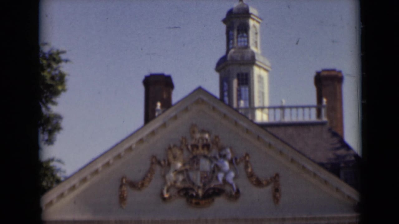1947 New York home-movie frame showing a historic gabled roof with gilded heraldry, brick chimneys, and a rooftop watch tower against blue sky.