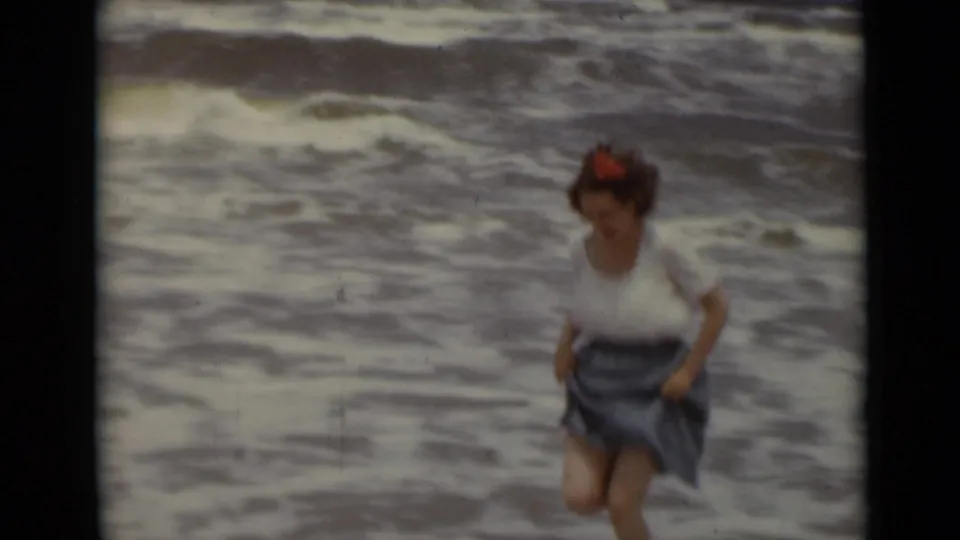 1947 home-movie beach frame showing a woman with a red hair flower, white top, and dark skirt moving through foamy surf.