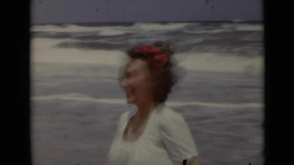 A smiling woman with red flowers in her hair appears in soft-focus 1947 New York beach footage with surf behind her.