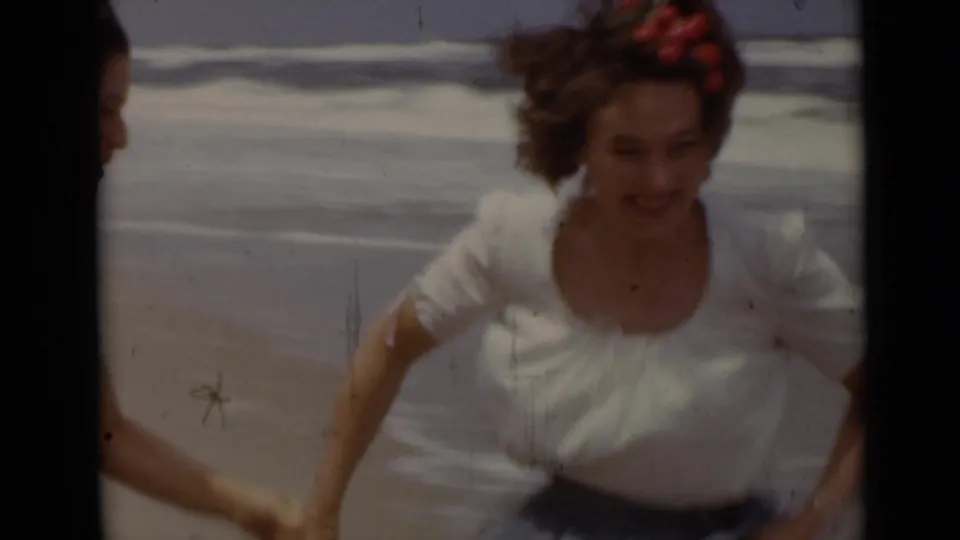 A woman in a white blouse and blue skirt runs toward the camera at the beach, holding another person's hand at the edge of frame.