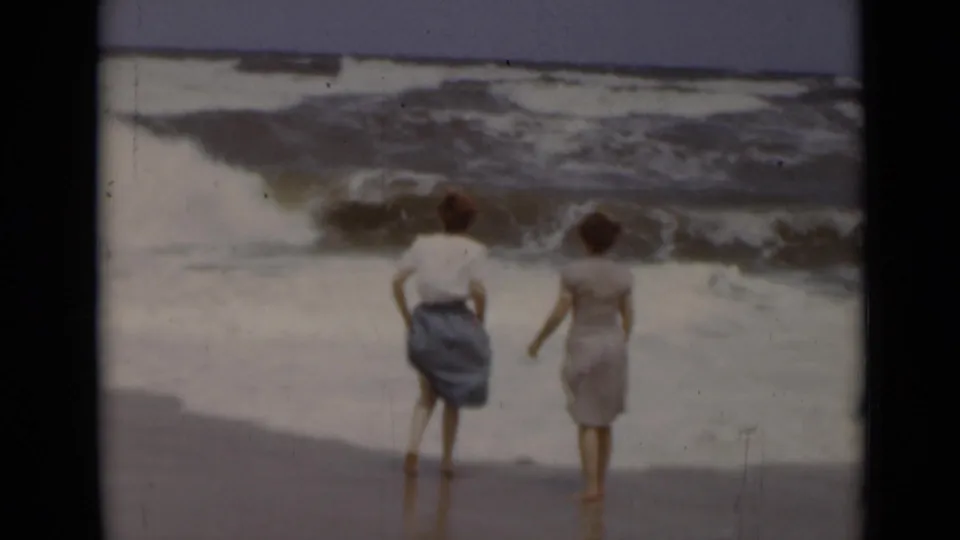 Two women in skirts stand ankle-deep near the surf in 1947 New York beach home-movie footage, seen from behind.