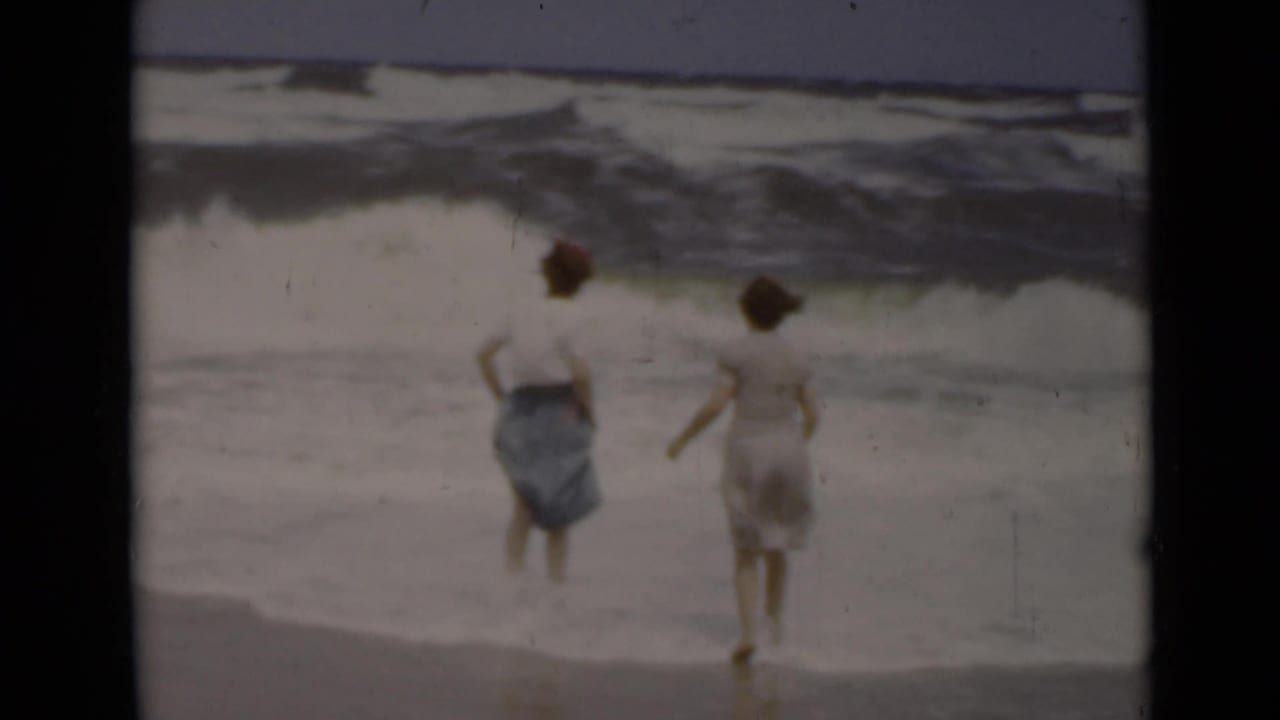 Two women stand near rough surf on a beach, seen from behind.