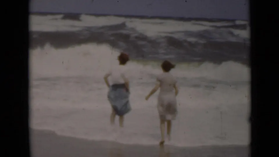 Two women in 1940s clothing stand at the waterline on a New York beach as heavy surf rolls in.