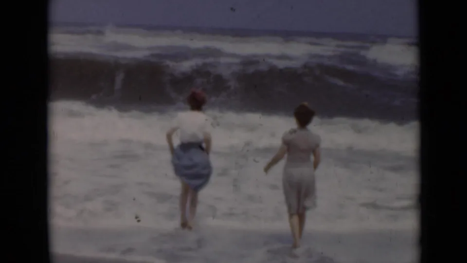 At a New York shoreline, two women are seen from behind near foaming waves in 1947 home-movie footage.