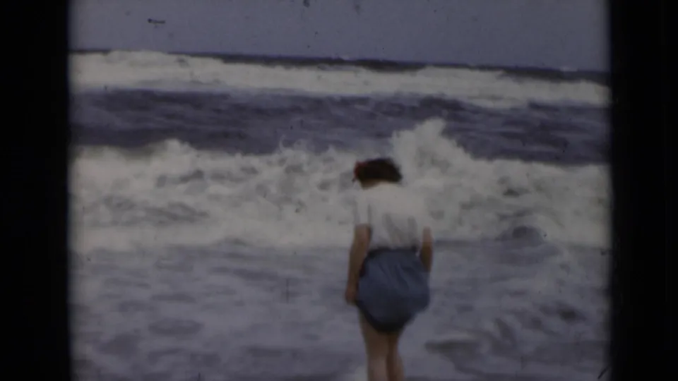 In supported 1947 New York home-movie footage, a woman faces the ocean as surf breaks near her feet.