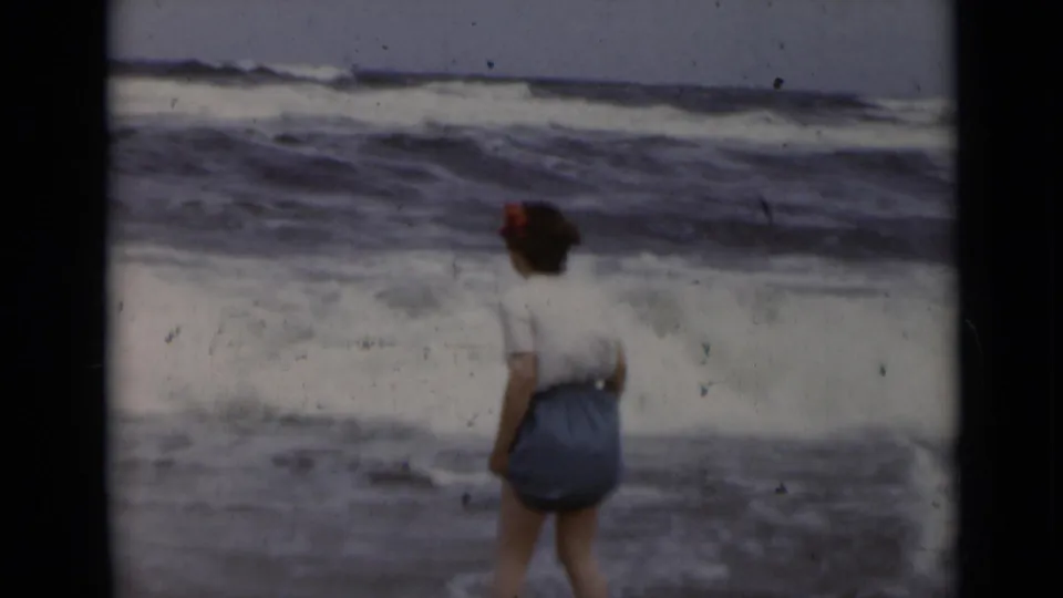 A woman in casual beach clothing faces rolling surf in 1947 New York archival home-movie footage.