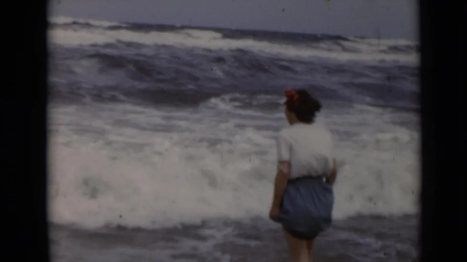The woman stands close to foamy waves, with rough ocean water filling the background in a 1940s beach scene.