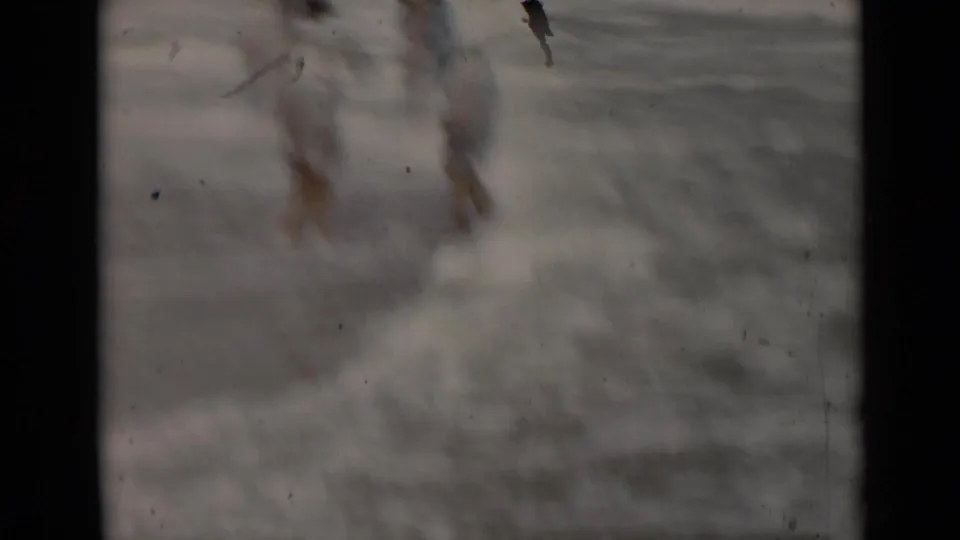 A soft, blurred 8mm frame shows the woman's lower body and feet near shallow surf at the shoreline.