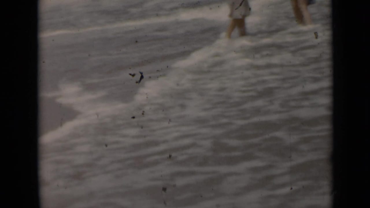 Two partial figures walk in shallow surf near the edge of the beach.