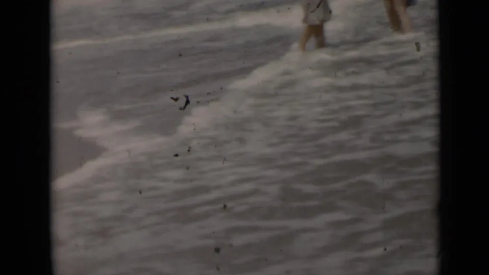 Soft 8mm frame from a 1947 New York beach scene showing partial adult figures walking through shallow shoreline water.