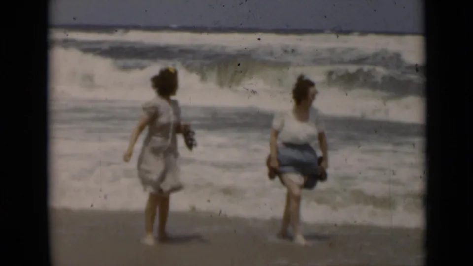 Two adult women walk barefoot along a New York shoreline in 1947, with foamy waves breaking behind them.