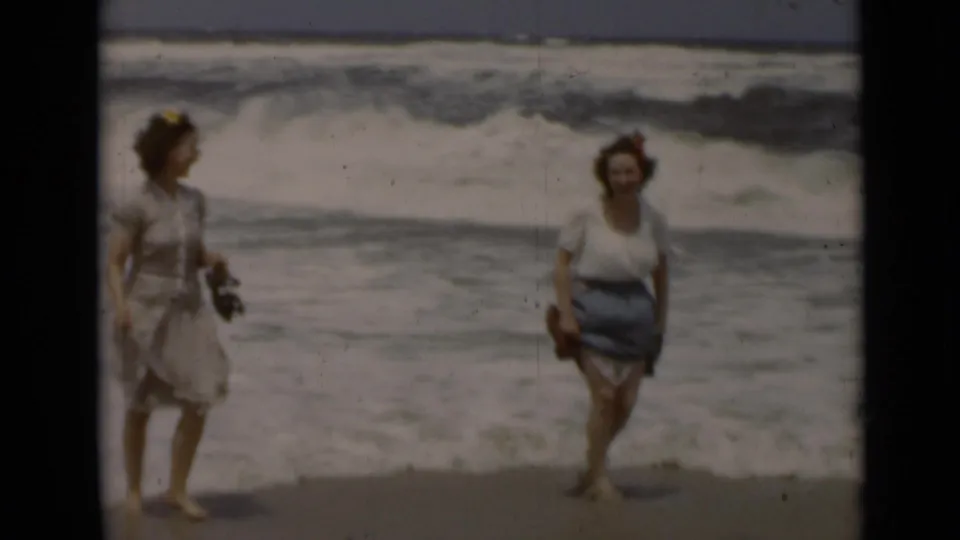 Two adult women in period beach clothing walk toward the camera on a 1947 New York beach as surf rolls in behind them.