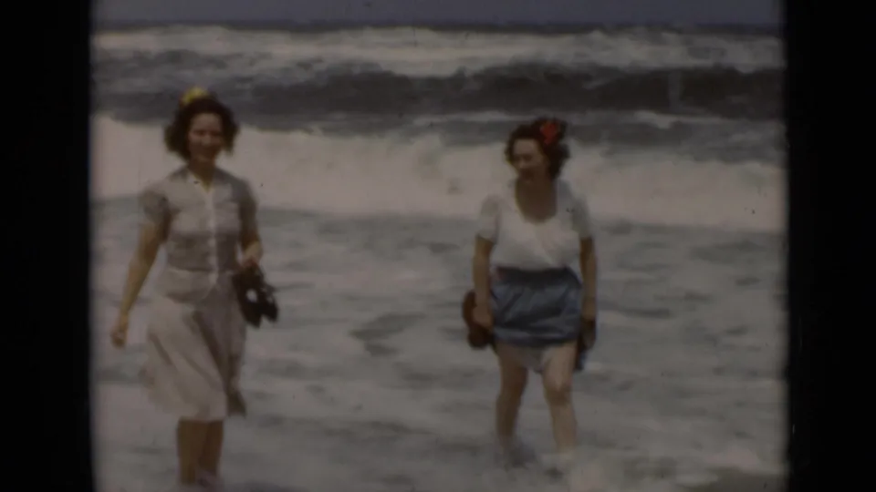 Two adult women are seen at the water’s edge on a 1947 New York beach, framed against foamy ocean waves.