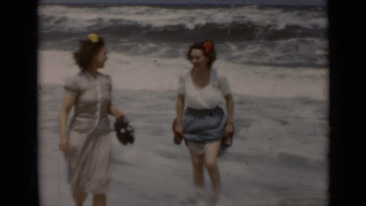 Two women walk barefoot near ocean waves, holding sandals on a beach.