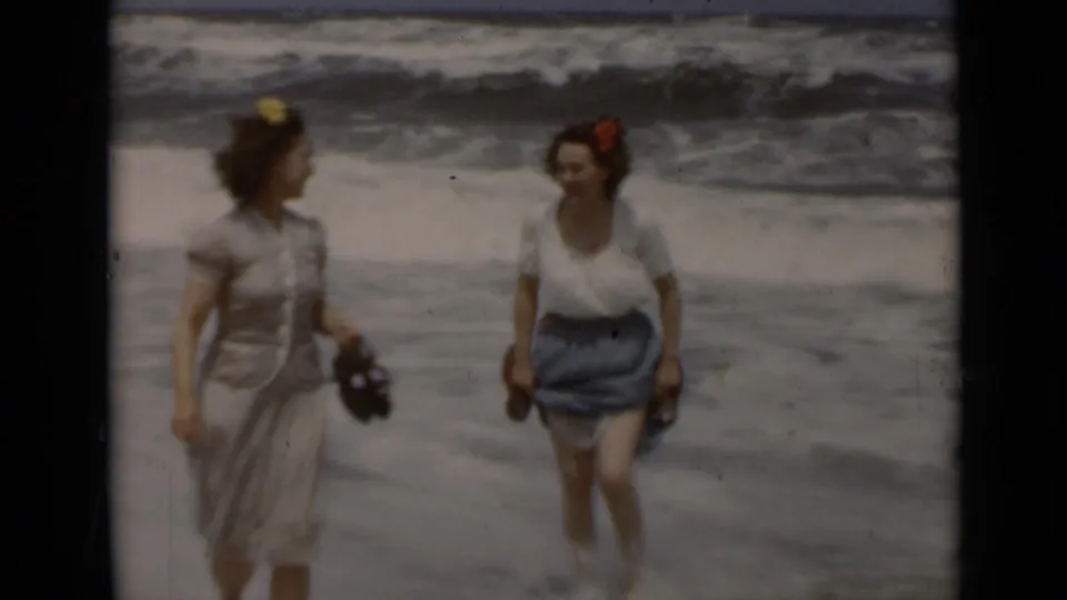 Two adult women in 1940s clothing walk along a New York beach, one in a light dress and one in a white blouse with blue skirt.