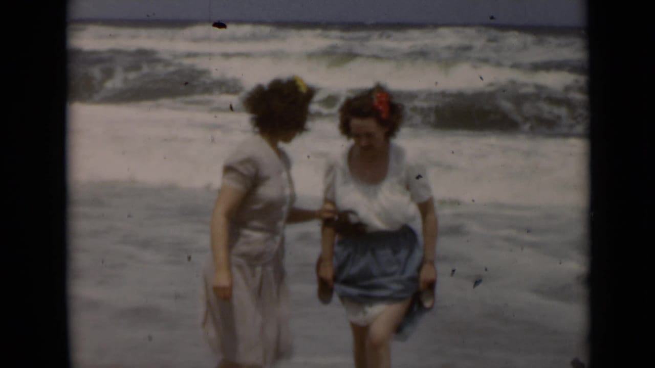 Two women stand close together at the shoreline while waves break behind them.