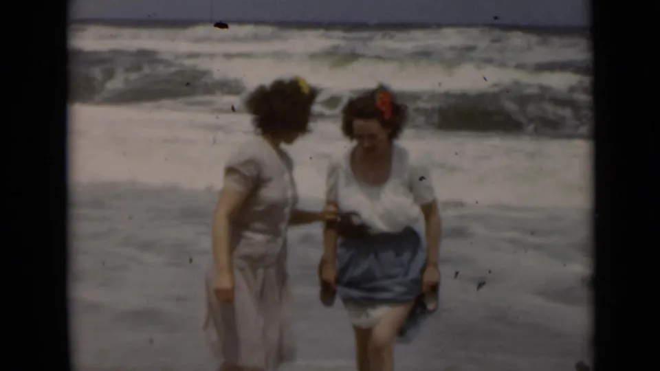 Two women on a New York beach appear to talk near shallow water, with flower hair accessories and sandals visible.