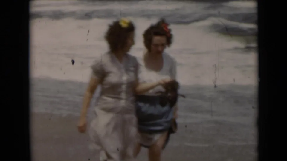 Two adult women in vintage beach attire walk together along the New York shoreline, partially blurred by 8mm motion and soft focus.
