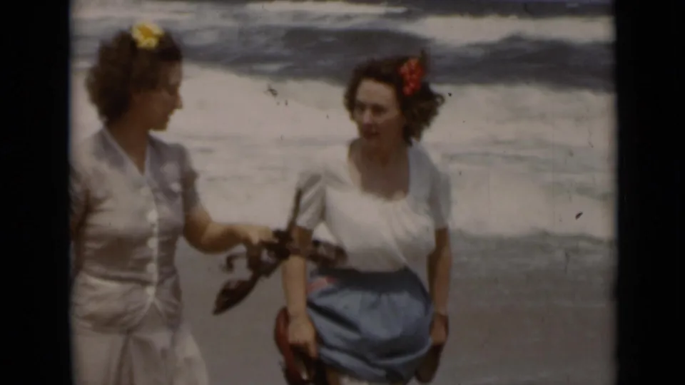Two women on a 1947 New York beach smile and converse, holding sandals as ocean waves break behind them.