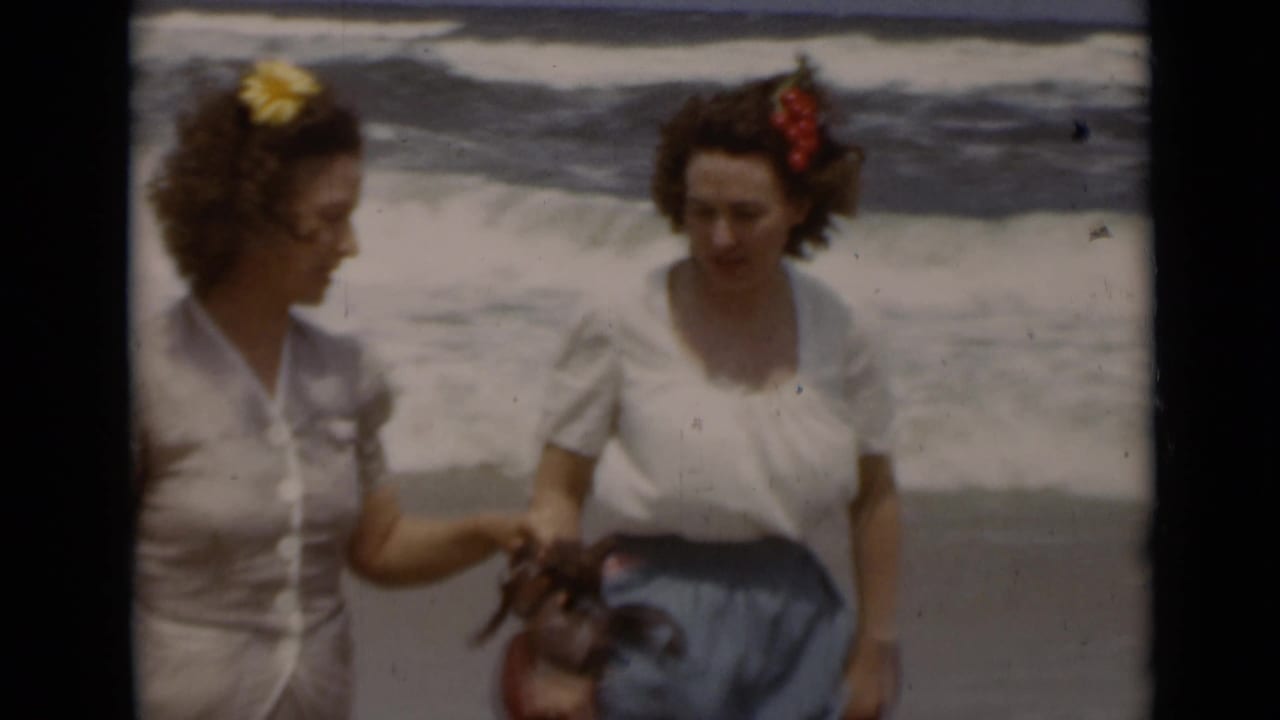 Two women walk on a beach with surf behind them, hair blowing in the wind.