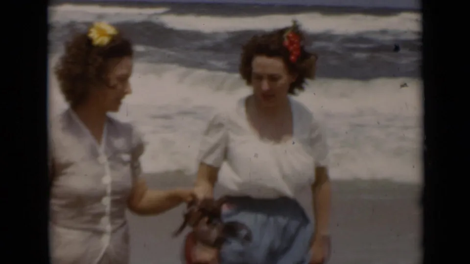 Two women in 1940s attire walk together on a New York beach, with white surf behind them and flowers in their hair.