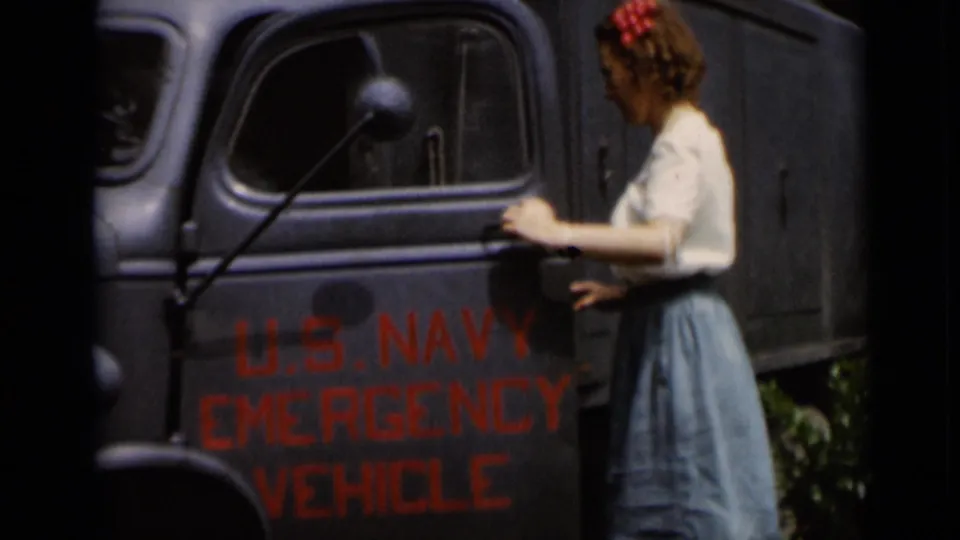 A woman in a white blouse and blue skirt stands beside a blue-gray U.S. Navy Emergency Vehicle with red lettering.
