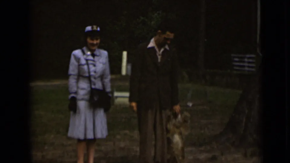 The couple remains in view with a small dog beside them, framed by trees and grass in the 1947 New York home-movie scene.