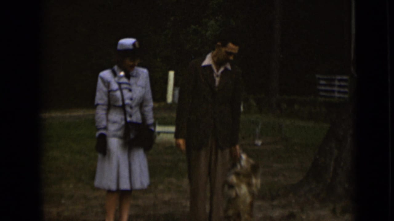 1947 New York home-movie frame showing a well-dressed couple walking a small dog in a grassy park area.