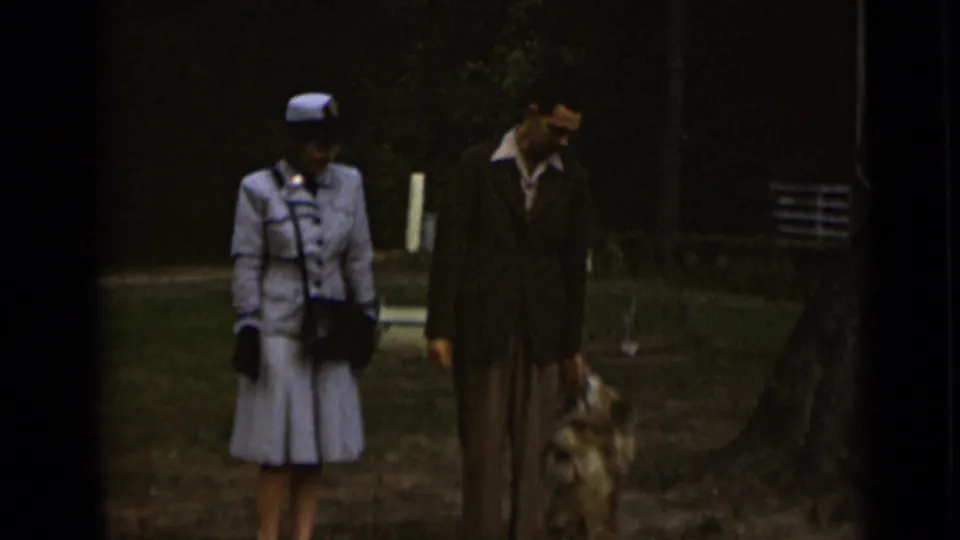 1947 New York home-movie frame showing a well-dressed couple walking a small dog in a grassy park area.