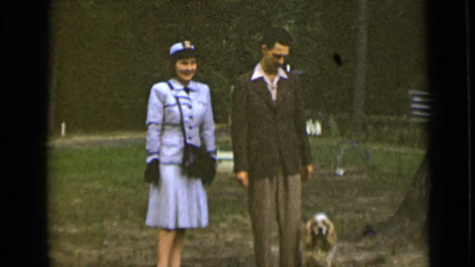 The woman in a pale suit smiles toward the camera while the man looks downward, with the dog low at the edge of the frame.