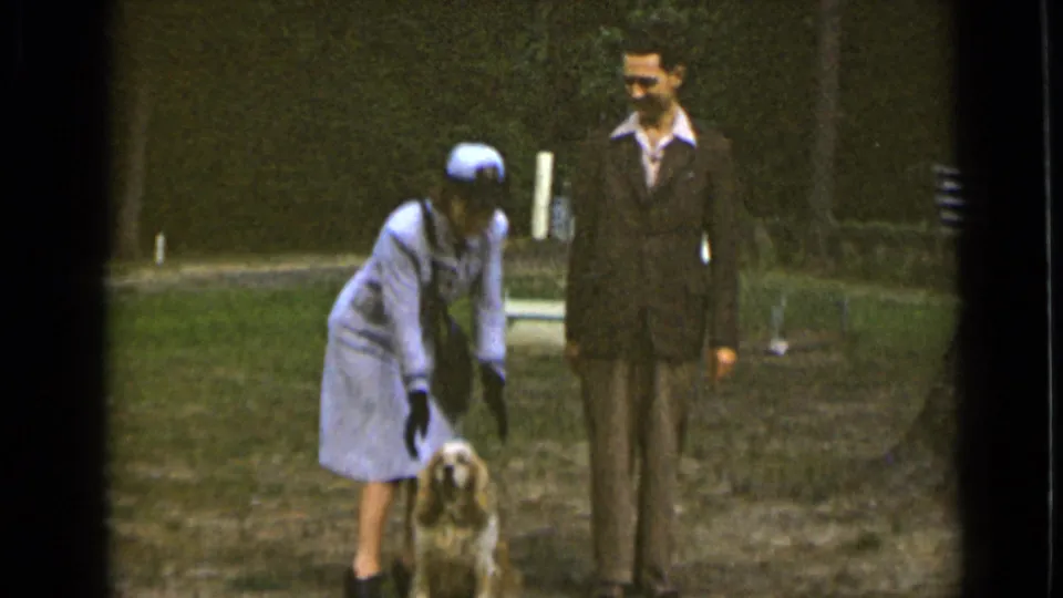 In the park footage, the uniformed woman bends down toward the leashed dog as the man in a dark suit looks on.