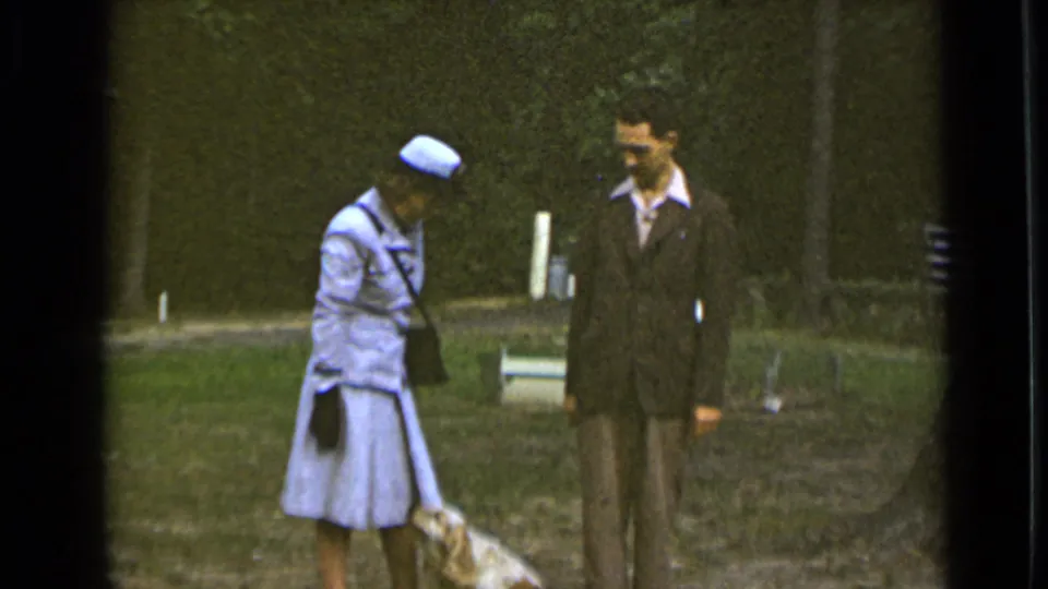 In the park, the woman stands upright beside the dog while the suited man faces her, with trees and a path behind them.